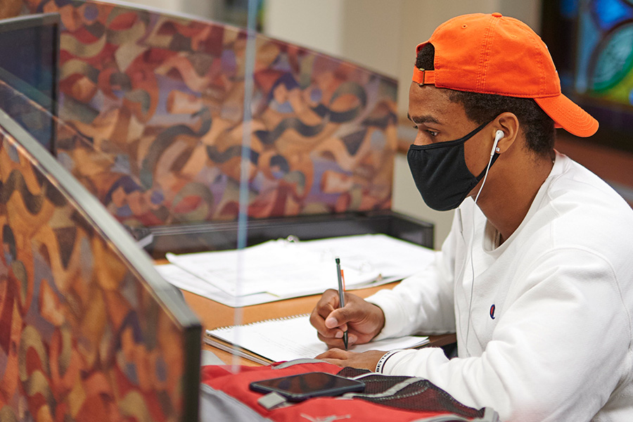 male student in an orange hat and white shirt, wearing a black mask, sitting at a desk, writing on paper and studying,