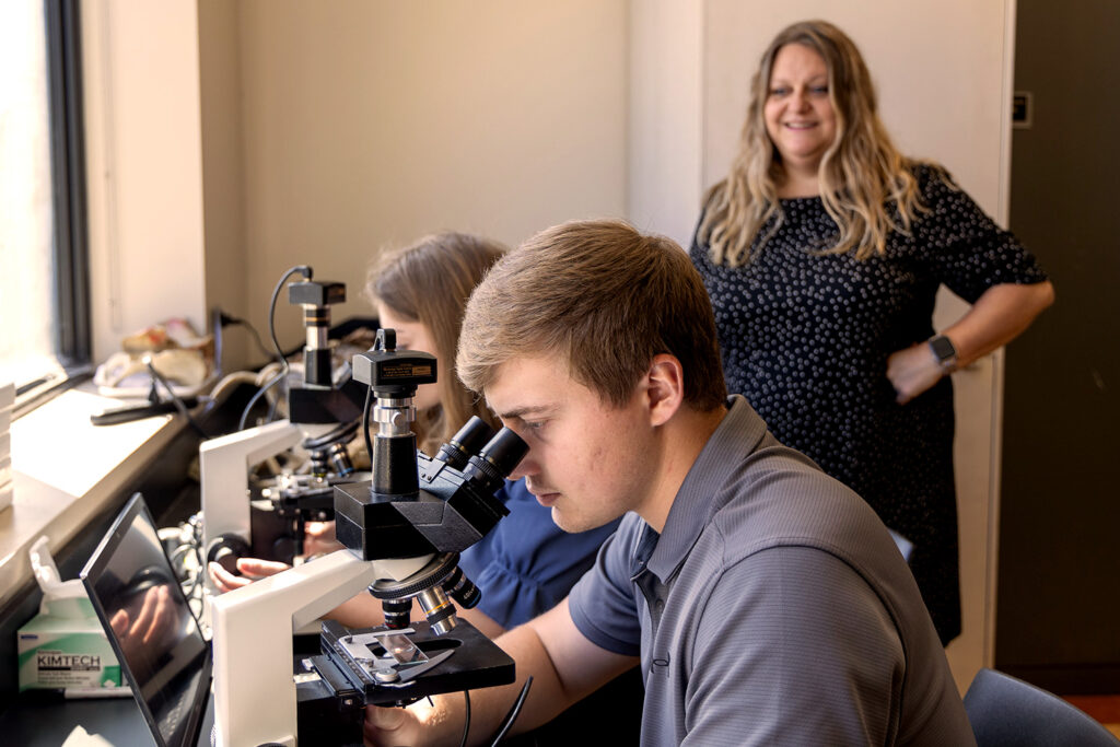 two students looking through microscopes while profefssor looks on